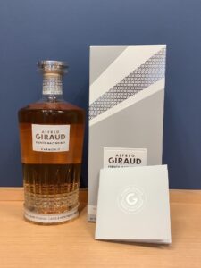FuFP7hOanNDb7FeaCBwqGB7dQrxCKPEuhSwUCTGw お酒買取専門店LIQUORJOYリカージョイ Bottle of Alfred Giraud French malt whisky Harmonie with a textured glass base placed next to its grey patterned box and brochures on a wooden surface against a blue wall