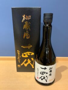 MHwSC3a0xEdS3oOzR77S9PYWCHRMfoZngvJIKaCQ お酒買取専門店LIQUORJOYリカージョイ A dark glass sake bottle with a white label and black cap standing beside a tall black box with gold Japanese calligraphy