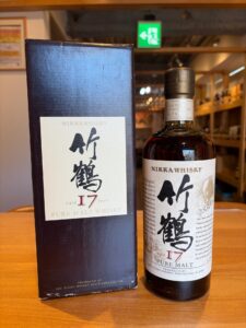 Bottle of Nikka Whisky 17 Year Pure Malt beside its dark box on a wooden table in a shop setting cartoon background blurred behind