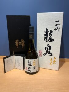 Japanese sake bottle with white label and black kanji flanked by a black gift box with gold emblem and a white box with bold calligraphy on a wooden surface