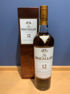 Bottle of The Macallan 12 Years Old Highland Single Malt Scotch Whisky with its white label beside the matching brown box against a blue wall