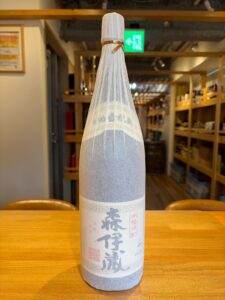 aVxKpH8B9YW7S4omMP9UNlpUYUFyPAjsh5KX2FC6 お酒買取専門店LIQUORJOYリカージョイ Frosted sake bottle on a wooden table inside a shop label with Japanese characters visible in the foreground