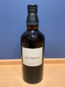 c6hKbJFy3uemZsfpW3owLEG4YfFNEOxFyarCuG2f お酒買取専門店LIQUORJOYリカージョイ Brown glass bottle with black cap and beige label reading Age Unknown on a wooden surface against a blue wall