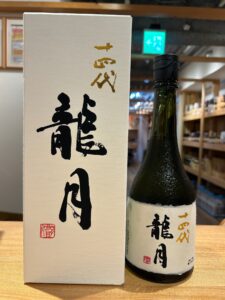 Sake bottle with a white textured box showing black and gold Japanese calligraphy and red seals set on a wooden surface in a store