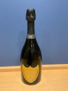 lMILnVI0oNVWvXf7RtD6sZZb8hOaRWTtMPJCNX4A お酒買取専門店LIQUORJOYリカージョイ Champagne bottle with a black foil neck and gold Dom Pérignon label on a wooden table against a blue wall