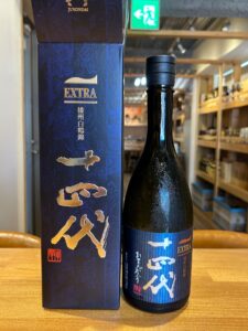 xwDnguJxLV1MMsLYbUtt5HzgbfmGe9dj4iNmLhc5 お酒買取専門店LIQUORJOYリカージョイ Dark glass bottle with a blue label that says EXTRA and gold Japanese characters placed beside a matching blue box on a wooden table in a shop