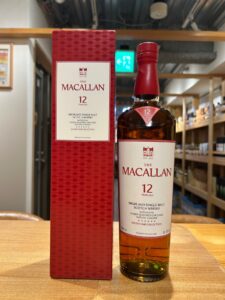 The Macallan 12 Year Old Highland Single Malt Scotch Whisky bottle beside its red patterned box on a wooden surface in a shop setting
