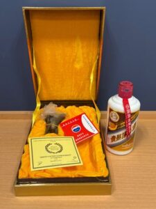 1 お酒買取専門店LIQUORJOYリカージョイ Open gift box lined in yellow containing a bottle of liquor with a red ribbon a yellow certificate and small cards on a golden fabric interior