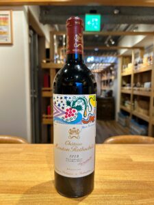 1 お酒買取専門店LIQUORJOYリカージョイ Bottle of Château Mouton Rothschild 2023 wine standing on a wooden table inside a wine shop with a colorful ornate label