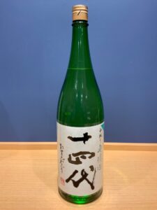 2uvcJPoyDKn5hjmy6xIzydj7AsMsfJVvh7bMvV8k お酒買取専門店LIQUORJOYリカージョイ Green glass sake bottle with a tan cap on a wooden table large white label with bold black kanji shapes in the center