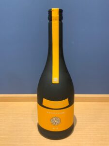 Dark bottle with a wide orange label and a vertical yellow stripe standing on a wooden table against a blue wall