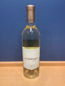 Tall white wine bottle with a foil neck sitting on a wooden surface against a blue wall label reads Sauvignon Blanc and estate bottled 2025 Napa Valley