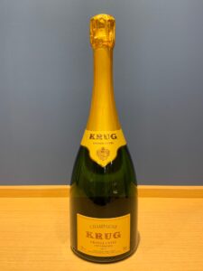 UGiOPNochqjw8nbPG5OKrVleTisCBV3YcJ5kYCsg お酒買取専門店LIQUORJOYリカージョイ Champagne bottle with gold foil label reads Krug Grande Cuvée on a wooden table against a gray wall