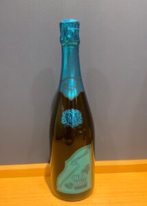 Bottle of Champagne with blue foil and turquoise label reading Soulmei or similar standing on a wooden surface against a gray wall