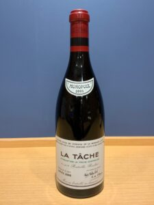 YfTFKVMjXQCfm2jOGXuKmGVuOYhX5ctYfoRbfrFN お酒買取専門店LIQUORJOYリカージョイ Wine bottle with red capsule and white label reading La Tâche 2005 on a wooden table against a blue wall