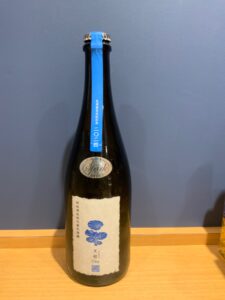 nTQHIliKYk5zcKVuBeN6TgZq1mrRuPdlpTalbxq9 お酒買取専門店LIQUORJOYリカージョイ Bottle of sake on a wooden surface brown glass with a blue tape seal along the neck and a white label featuring blue floral designs