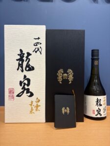 Gift style Japanese sake set white textured box with black calligraphy a black box with gold crest and a bottle with white label on a wooden surface