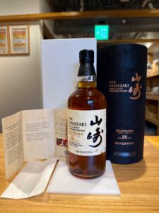 1 お酒買取専門店LIQUORJOYリカージョイ Bottle of The Yamazaki Mizunara Japanese whisky in a dark blue cylindrical box on a wooden table with a folded pamphlet nearby 2024 edition