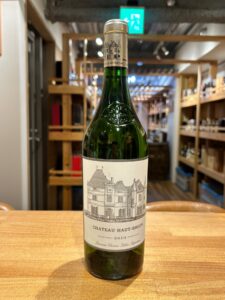1 お酒買取専門店LIQUORJOYリカージョイ Green wine bottle on a wooden counter in a shop label reads Château Haut Brion 2019 with château illustration