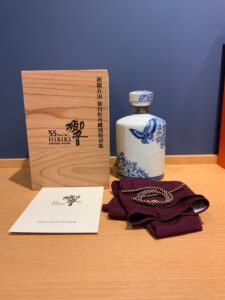 6cvoEOhU8T3zCIPhUxT0HH0yfeEHmwiNjaK4hZqo お酒買取専門店LIQUORJOYリカージョイ Wooden display box labeled 35 Years Old Hibiki Suntory Whisky beside a white and blue porcelain bottle with butterfly and floral patterns on a wooden surface against a blue wall