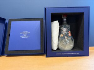 73YraHzWscfz4u6nWCBCYdMGHGNj3DM2GJuqL1f7 お酒買取専門店LIQUORJOYリカージョイ Opened blue gift box showing a decorative bottle of Suntory Blended Japanese Whisky 2025 with a white wrapping on a wooden surface