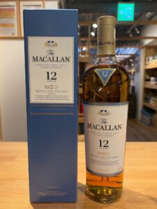 The Macallan 12 year old Highland Single Malt Scotch Whisky bottle beside its blue box on a wooden counter in a shop The label reads Triple Cask Matured and Natural Colour