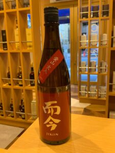 Tall brown sake bottle with red label and white kanji on a wooden counter inside a liquor store shelves of bottles visible in the background