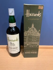 Bottle of Harrods Scotch Whisky with white label and white cap placed next to a dark Harrods of Knightsbridge box on a wooden surface