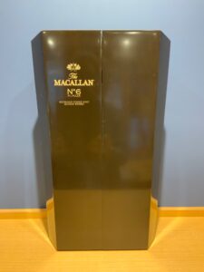 The Macallan No 6 Highland Single Malt Scotch Whisky gift box in dark brown with gold lettering positioned against a blue wall