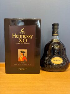 Box and bottle of Hennessy XO Extra Old Cognac on a wooden surface against a gray wall showing the brand name prominently