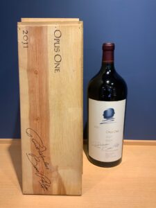 Wooden wine crate labeled Opus One 2011 beside a dark Opus One wine bottle on a light wood surface against a blue background