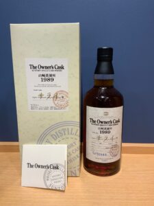 Bottle of The Owners Cask whisky with a cream box and paperwork on a wooden table against a blue wall showing the 1989 label