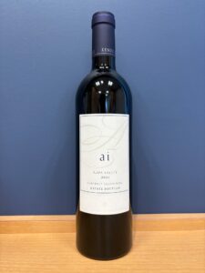 Full bottle of red wine on a wooden surface against a blue wall label reads Napa Valley 2021 Cabernet Sauvignon Estate Bottled