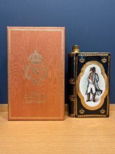 Two vintage perfume boxes on a wooden surface a tall light brown box with a crest and gold border and a black ornate bottle with a gold trim and an oval portrait of a man in period dress
