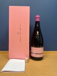 Pink wine box and pink labeled bottle with pink foil on a wooden surface beside an open booklet and a cork