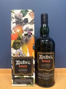Bottle of Ardbeg Dolce whisky beside its colorful box on a wooden surface against a blue wall with the labels readable as Ardbeg Dolce