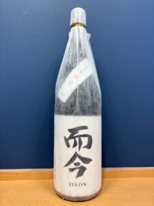 tmxLymZIppc4AnUCoPgSb7xFX8WFtiTYQeKfBS9I お酒買取専門店LIQUORJOYリカージョイ Wrapped sake bottle with white paper and a gold neck band labeled JIKON on a wooden table against a blue wall