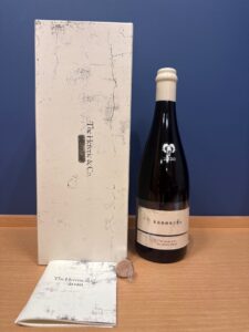 Wine bottle with a white wax seal stands next to a tall distressed white box and a folded booklet labeled The Heretic  Co 2020 on a wooden surface
