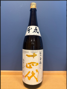 图片2026101915062178 お酒買取専門店LIQUORJOYリカージョイ Tall bottle of Japanese sake with white labels and large gold kanji characters standing on a wooden surface against a blue wall